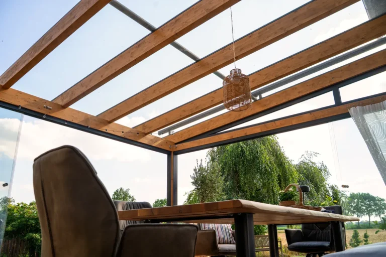 Blick von unten auf eine Terrassenüberdachung mit Glasdach und Holzsparren, darunter Esstisch mit Polsterstühlen und eine Rattan-Hängeleuchte, im Hintergrund Bäume und Felder.